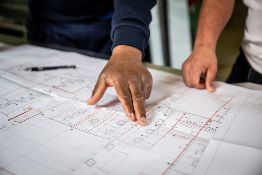 Hand wijst naar bouwtekening op tafel.